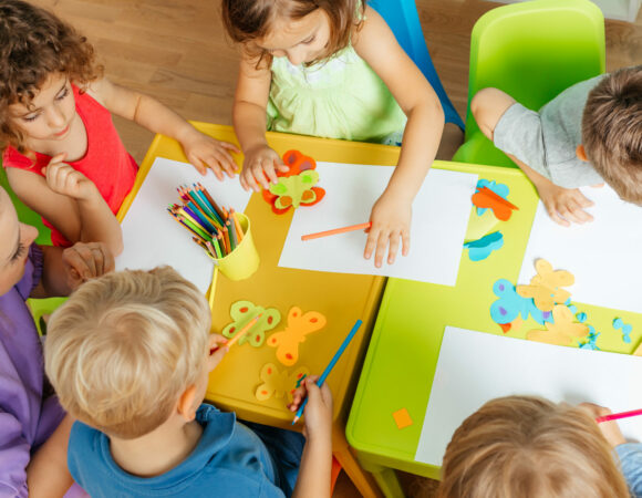 Top view kindergarten children during art lesson Art lesson at the kindergarten, top view. Lovely children glueing and cutting paper on colorful plastic tables. Children development concept.